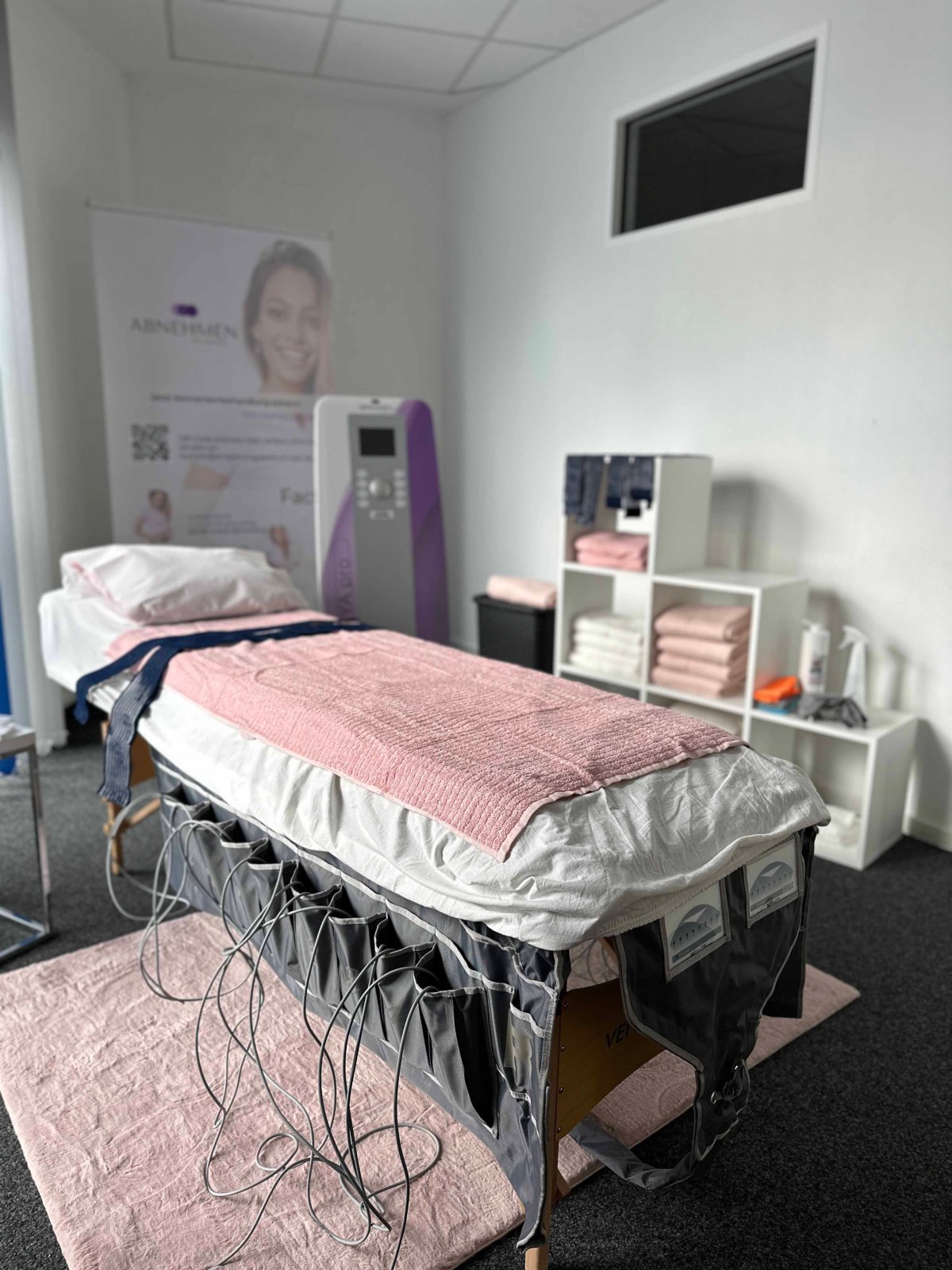 Behandlungsraum mit Liege, rosa Decke und MVA.pro-Gerät vor einem Regal mit Handtüchern